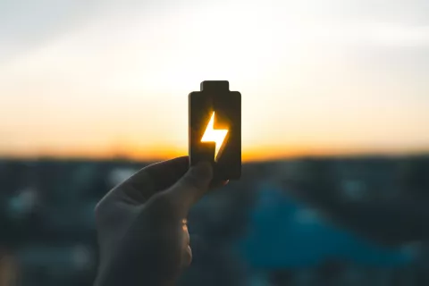 Eine Hand hält dem Umriss einer Batterie in den Himmel, während die untergehende Sonne durch das Strom-Symbol scheint und leuchtet.